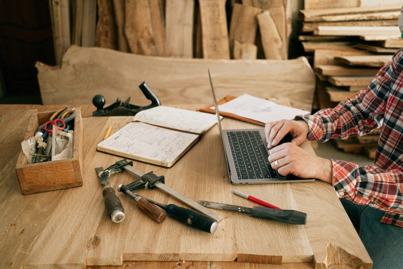 A carpenter using a laptop surrounded by tools in a wooden workshop, merging craft with technology.